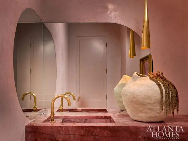 Pink textured walls and a large arched mirror above a pink marble vanity with double brass faucets and two textured vases.