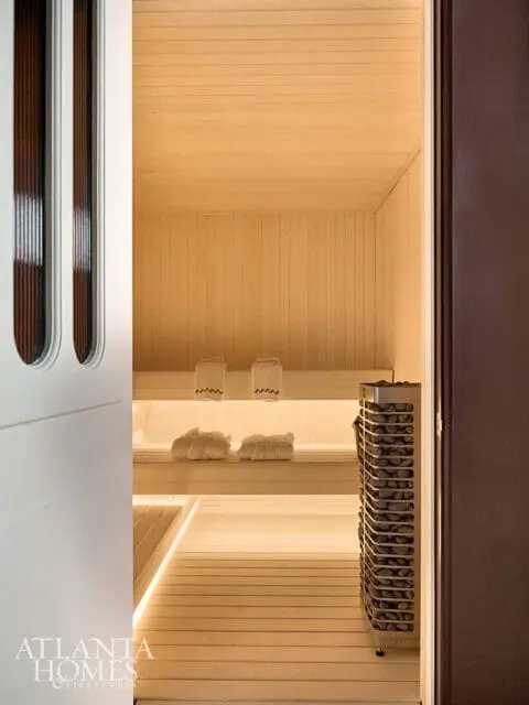 Modern sauna interior with light wood paneling, two-tiered benches, white towels, and a stainless steel sauna heater filled with stones.