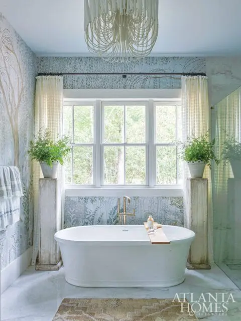 Freestanding white bathtub with brass faucet sits below a large window with sheer curtains. Botanical wallpaper and a beaded chandelier adorn the room.