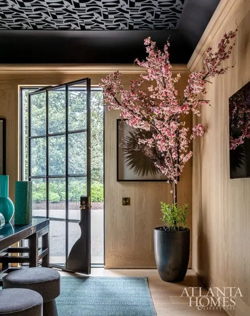 Interior entryway with light wood paneled walls, a black ceiling with a patterned design, a large potted cherry blossom tree, and a black metal-framed door opening to an exterior garden.