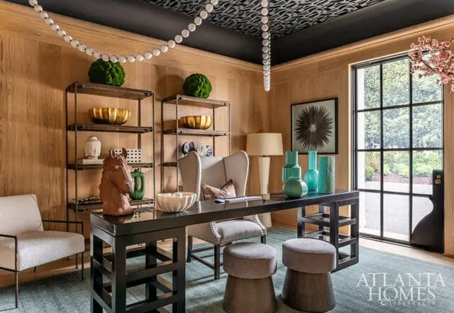 Wood-paneled office with a black desk, wingback chair, and two stools. Shelving holds decorative items, and a large window looks out onto a garden.