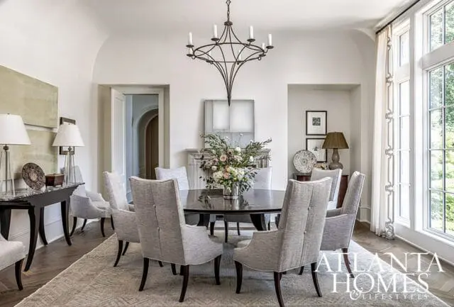 Dining room with a dark wood round table surrounded by upholstered chairs, a floral centerpiece, and a wrought iron chandelier.