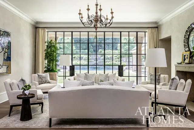 Living room with neutral-toned sofas and armchairs, a large grid window overlooking a garden, and a dark metal chandelier.