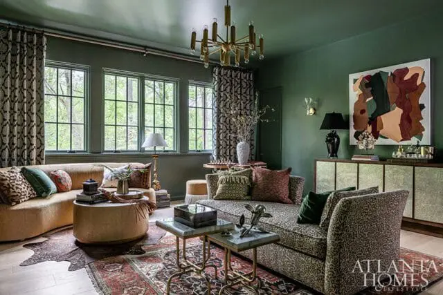 Dark green living room with a curved camel-colored sofa, a leopard-print chaise, patterned curtains, a large abstract artwork, and a brass chandelier.