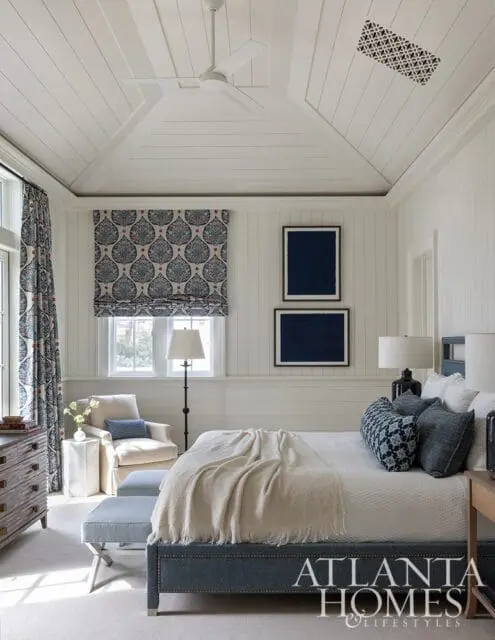 Bedroom with white paneled walls and ceiling, a white ceiling fan, blue patterned curtains and roman shades, a cream armchair, a blue upholstered bed, and two framed navy blue artworks.
