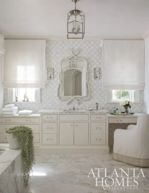 White bathroom with a double vanity, marble countertops, ornate mirror, patterned wallpaper, and a plush armchair.