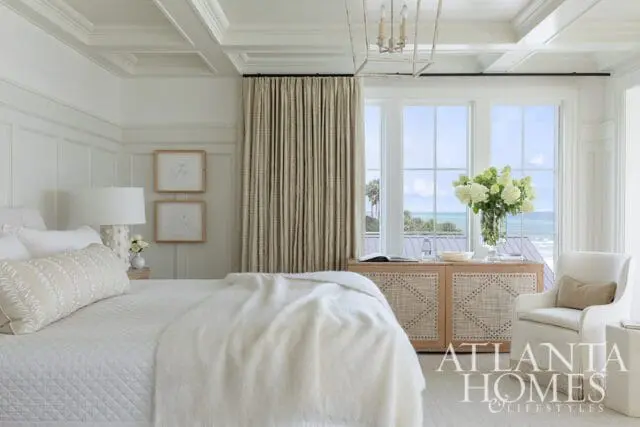 Primary bedroom with coffered ceiling, wainscoting, and large windows overlooking the ocean. Features a white quilted bed with a textured throw, a cane-front dresser, and a white armchair.
