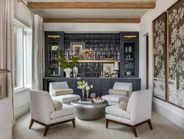 Living room with four light gray armchairs arranged around a silver coffee table, facing a dark gray built-in bar with black tile backsplash and glass-front cabinets.