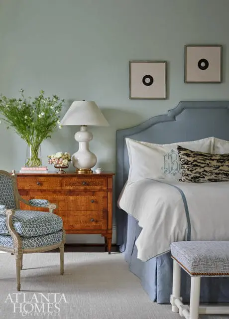 Bedroom with blue headboard, white bedding, wooden dresser, and patterned chair.