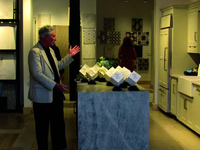 Man in blazer gesturing near marble island with awards and tile samples on wall