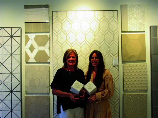 Two women holding awards in front of a wall of decorative tiles.