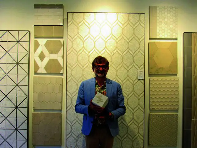 Man in blue jacket holding award in front of patterned wallpaper and tile samples