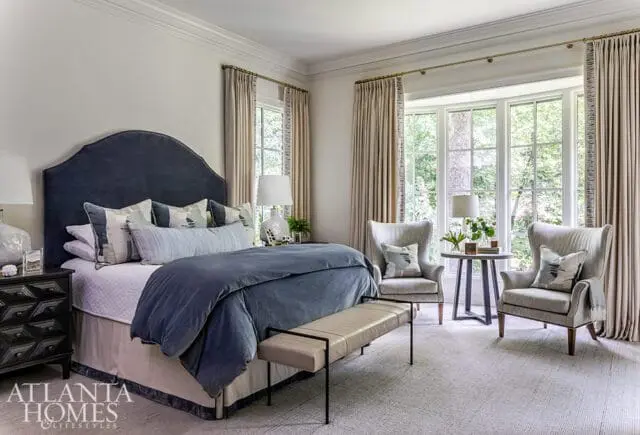 Bedroom with navy upholstered headboard, blue bedding, beige curtains, and two wingback chairs.