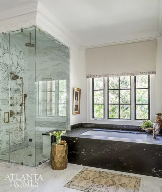 Marble shower with brass fixtures next to a black marble bathtub and window.