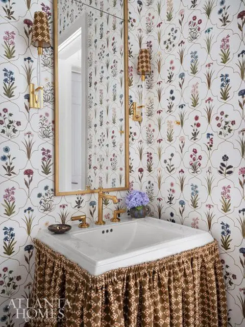 Powder room with floral wallpaper, gold faucet, and skirted pedestal sink.