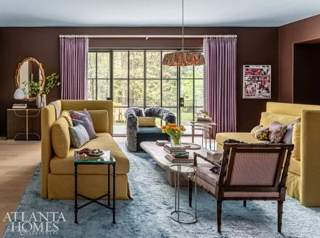 Living room with yellow sofas, plush armchair, and striped bench. Large window overlooks trees.