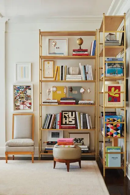 Gold bookshelves filled with books, art, and decor. A chair and ottoman are nearby.