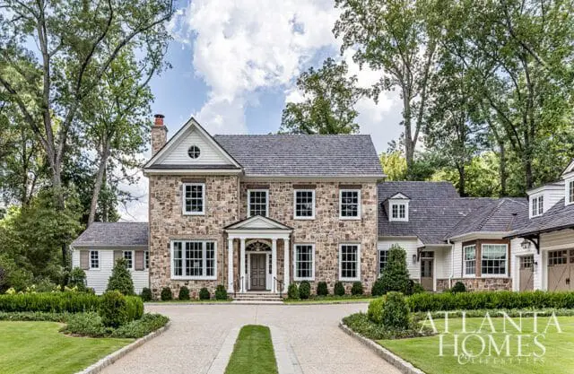 Stone and white house with gray roof, surrounded by trees and manicured lawn.