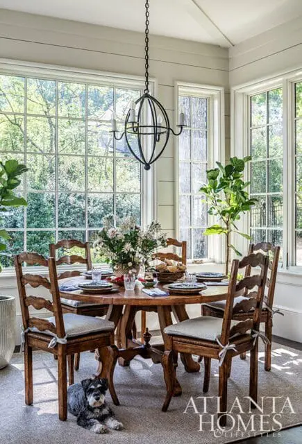 Dining room with round wooden table, chairs, and large windows overlooking greenery.