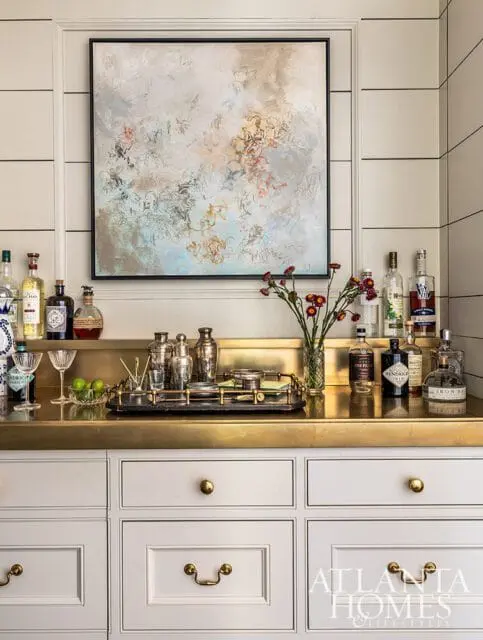 Bar area with brass countertop, white cabinetry, abstract art, and liquor bottles.