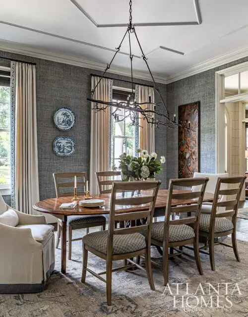 Dining room with oval wood table, upholstered chairs, textured gray walls, and ornate chandelier.