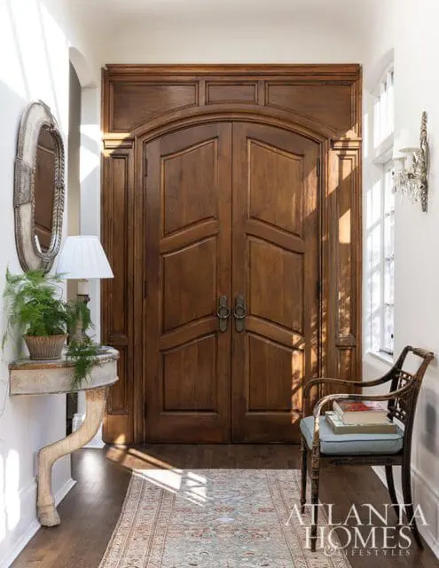 Foyer with arched double wooden doors, ornate mirror, console table, and wicker chair.