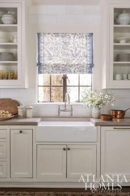 White kitchen with a floral Roman shade, farmhouse sink, and glass-front cabinets.
