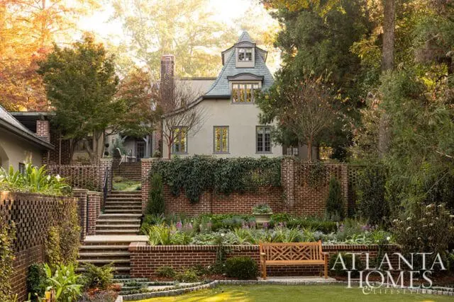 French-style house with gray slate roof, brick accents, and tiered garden with bench.