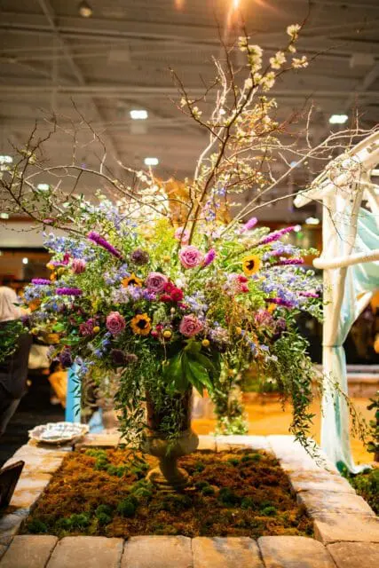 Large floral arrangement with roses, sunflowers, and branches in a stone urn on mossy ground