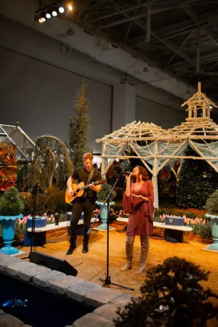 Musicians perform on a stage with a gazebo and floral displays.