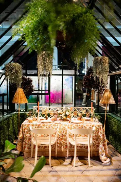 Greenhouse dining area with a floral tablecloth, white chairs, and hanging plants.
