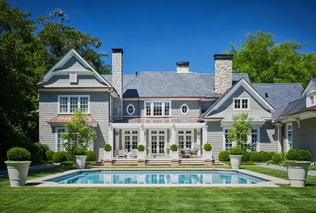 Shingle-clad house with stone chimneys and a blue slate roof, featuring a covered patio overlooking a swimming pool and manicured lawn.