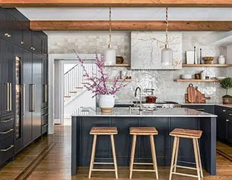 Dark gray kitchen island with three wooden stools, white tile backsplash, and wooden beams overhead.