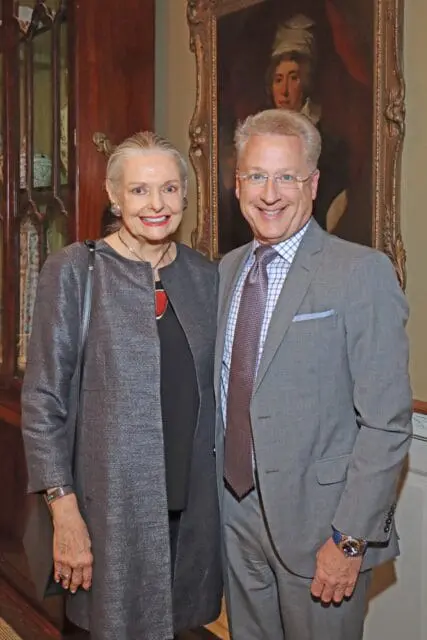 Woman in gray jacket and man in gray suit stand before a painting.