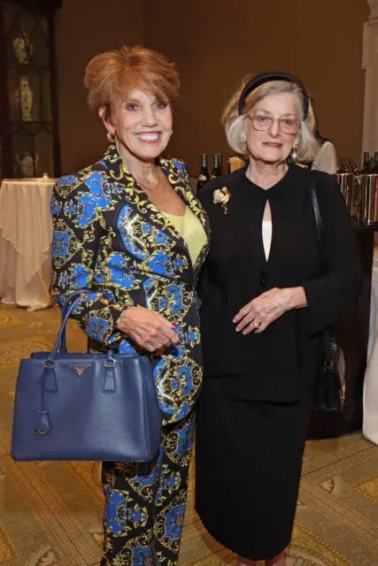 Woman in patterned suit with blue handbag and woman in black suit with brooch