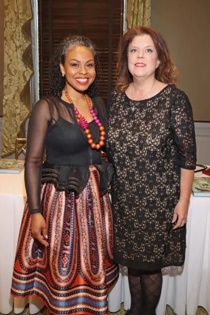 Two women posing for a photo. The woman on the left wears a black mesh jacket and a colorful