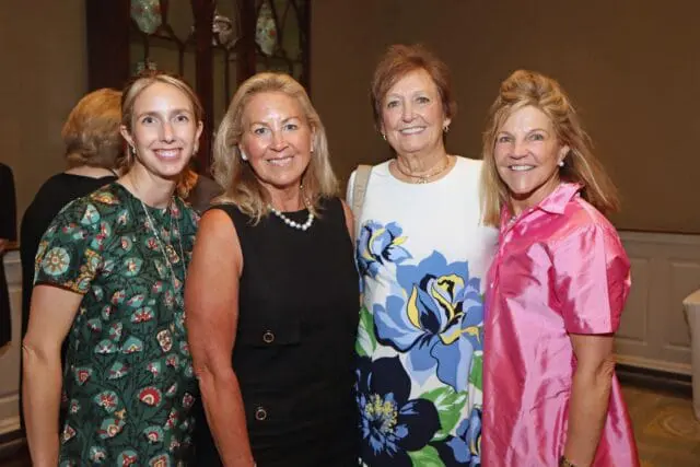 Four women smiling, one in a green floral dress, one in black, one in a white floral dress, and one