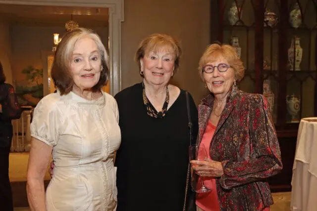 Three women posing for a photo. The woman on the left wears a cream dress. The woman in the middle