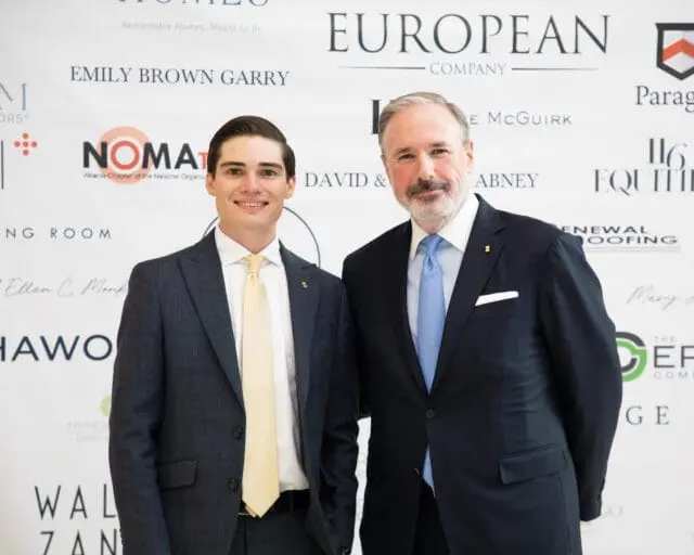 Two men in suits stand in front of a backdrop with sponsor logos.