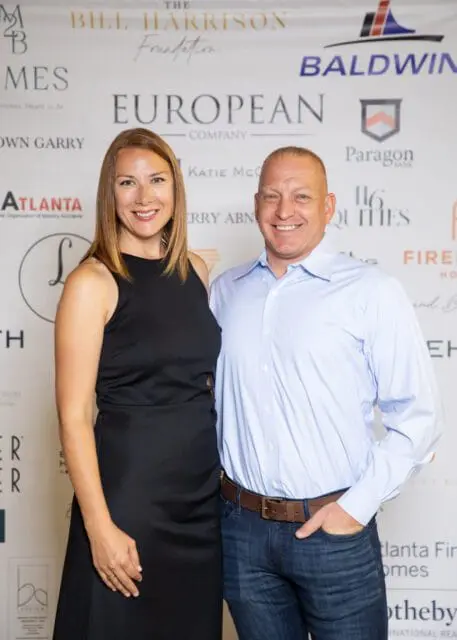 Man in blue dress shirt and woman in black dress smiling, standing in front of a sponsor banner