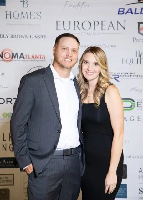 Man in a pinstripe suit and woman in a black dress smiling in front of a backdrop with sponsor
