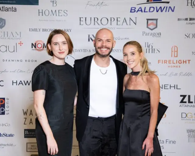Three people in black formal wear pose in front of a backdrop with sponsor logos.