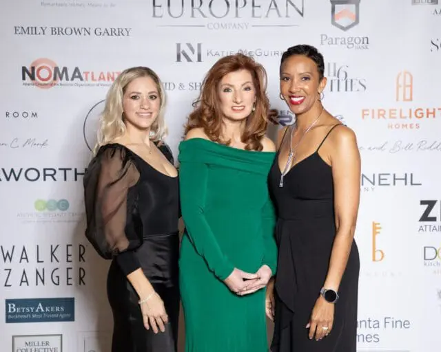Three women posing in front of a sponsor wall at an event.