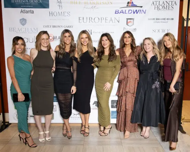 Eight women in formal dresses pose for a photo in front of a sponsor backdrop.