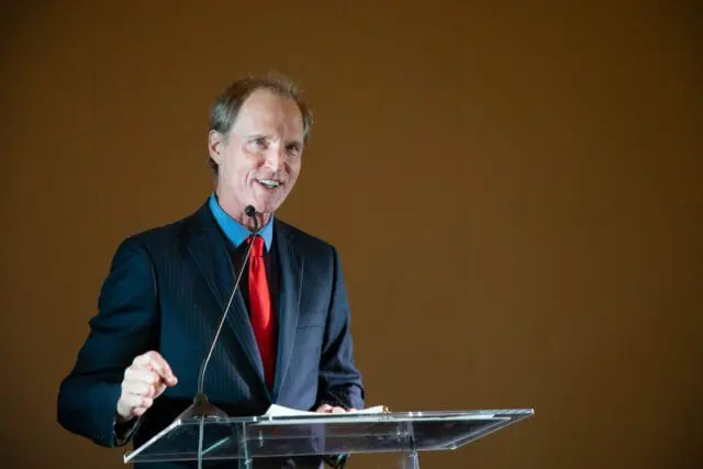 Man in a dark pinstripe suit and red tie speaking at a podium.