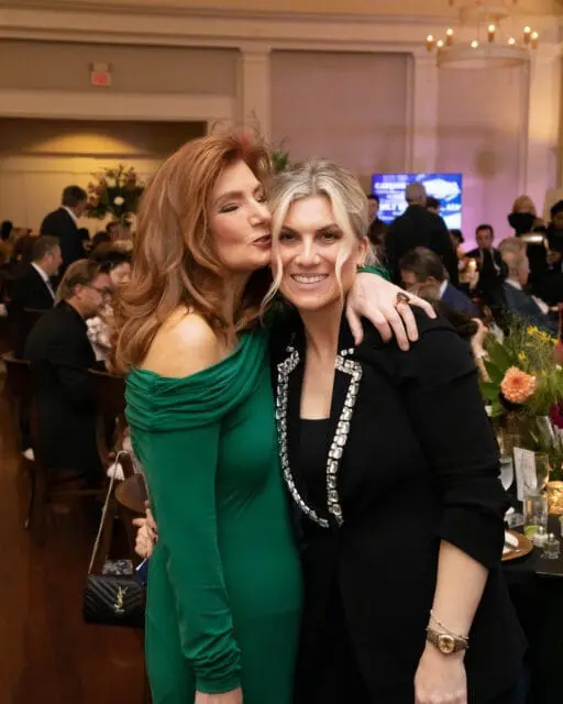 Woman in green dress whispers to woman in black jacket with jeweled collar.