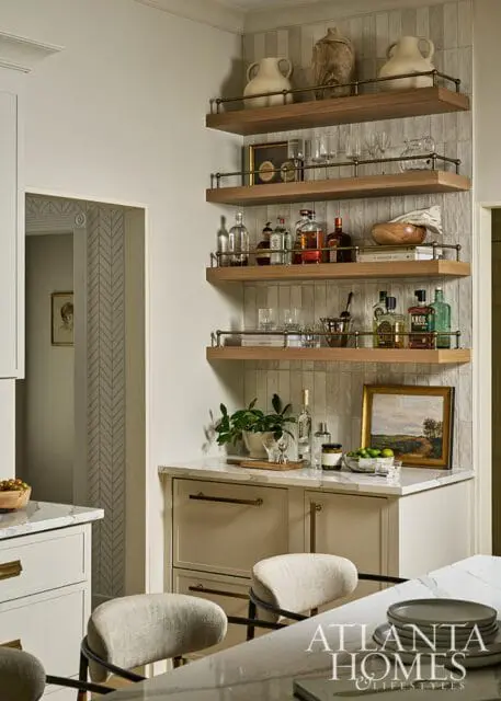 Bar area with wood shelves holding bottles and glassware, marble countertop, and neutral stools.