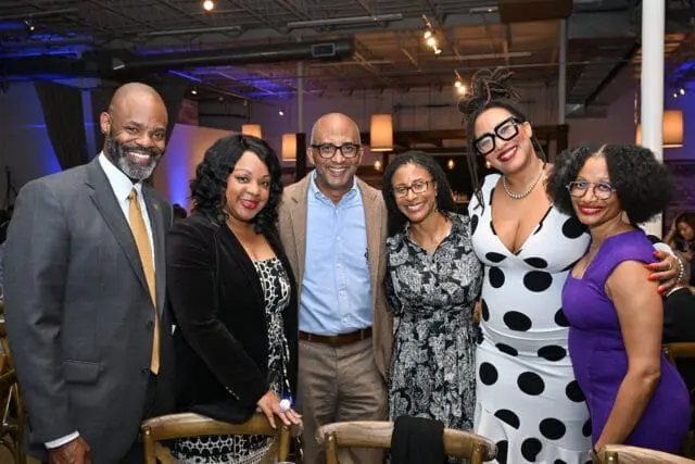 Six people smiling at a formal event. A man in a suit and gold tie stands left.