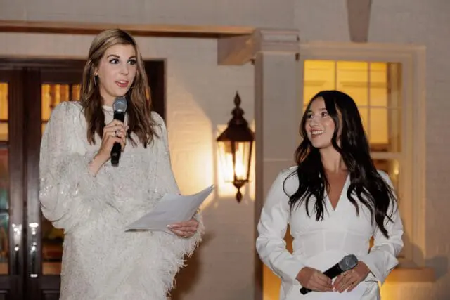 Two women in white dresses holding microphones and papers.