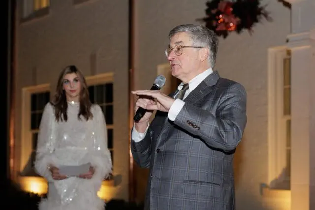 Man in plaid suit speaking into microphone, woman in sequined dress behind him
